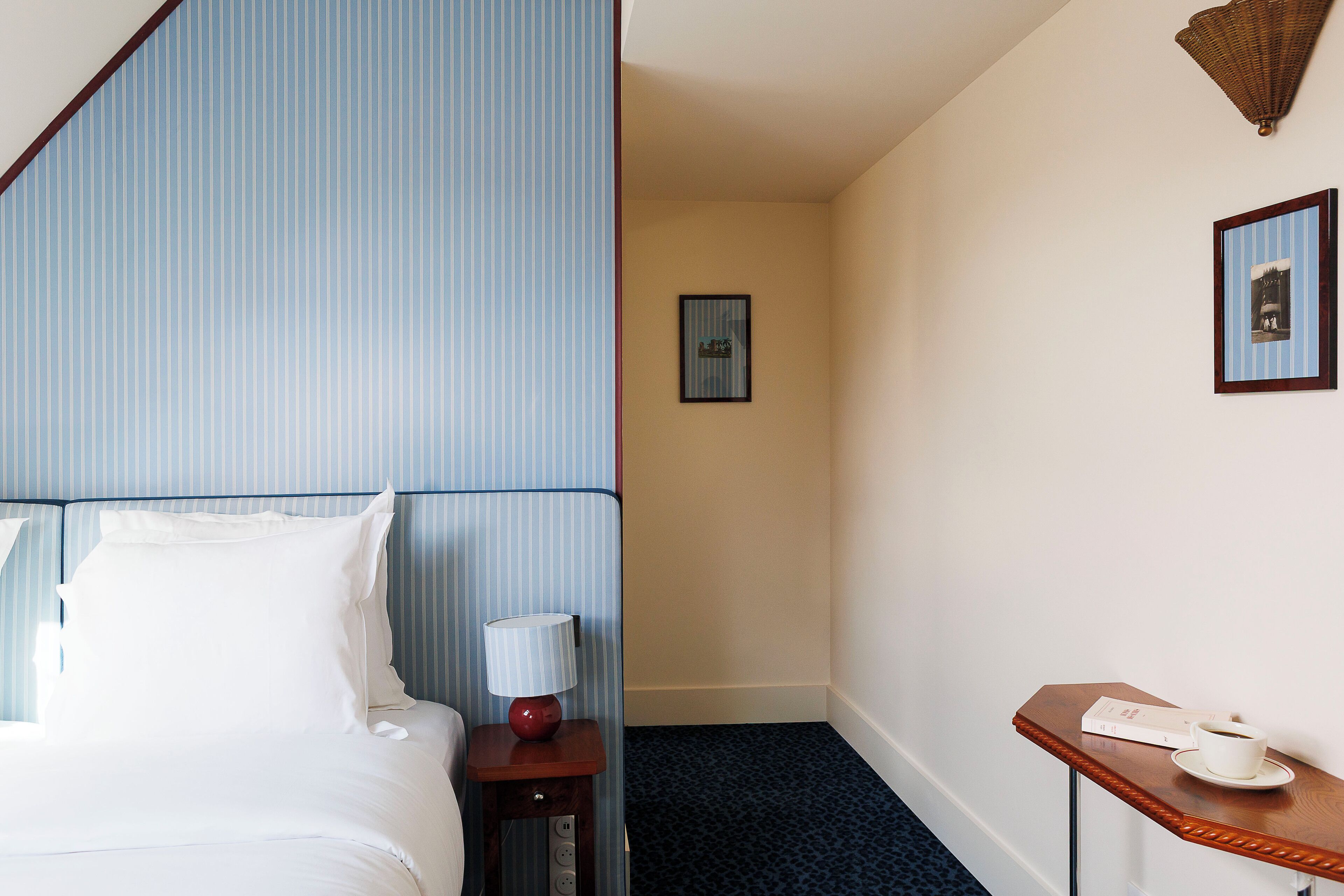 A hotel room with a bed, a bedside table with a lamp, a small table with a cup and a menu, and a framed picture on the wall.