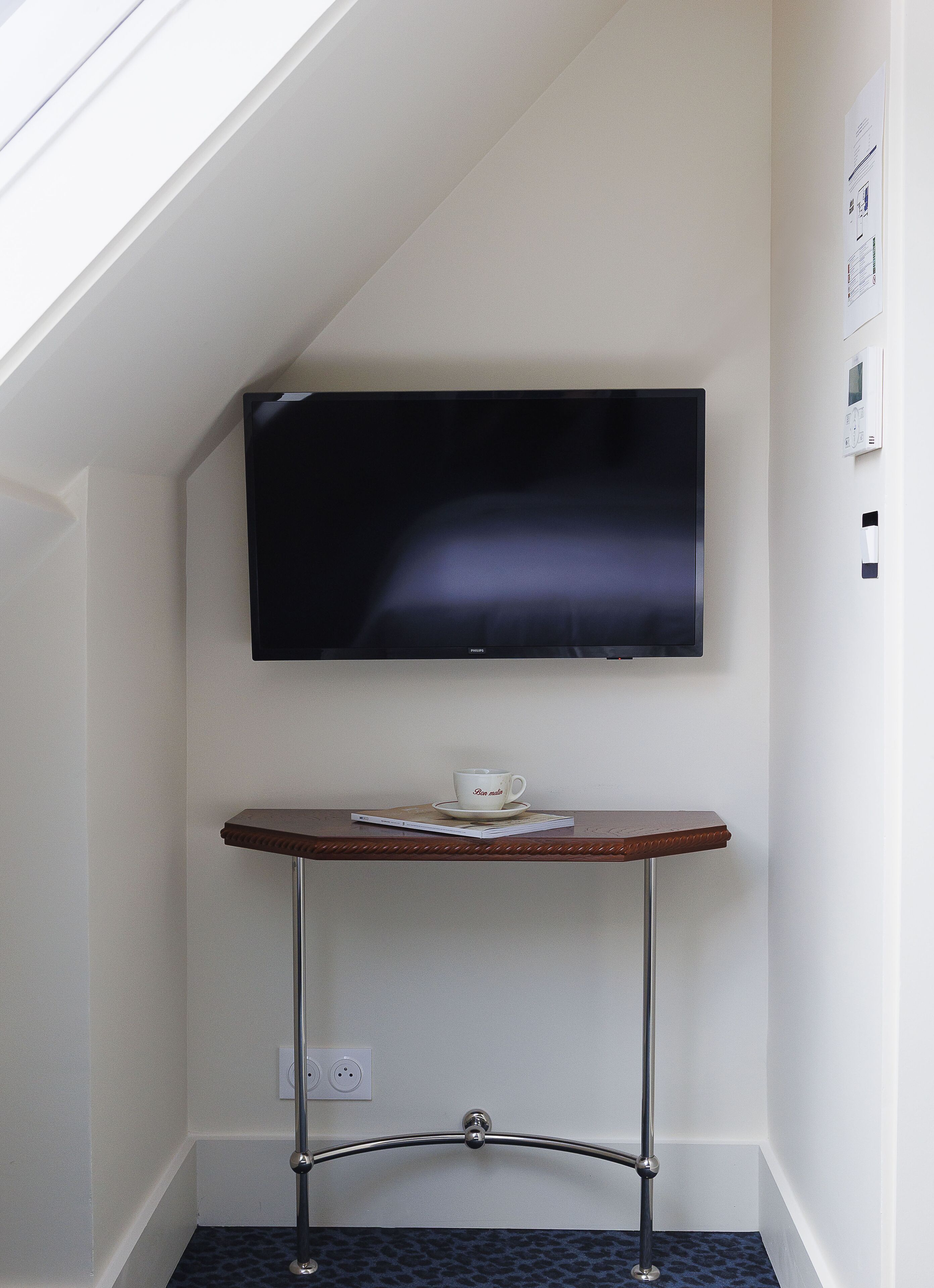 Ein Eckzimmer mit Flachbildfernseher an der Wand, einem kleinen Holztisch mit Tasse und Untertasse sowie einem Hocker mit Metallgestell.