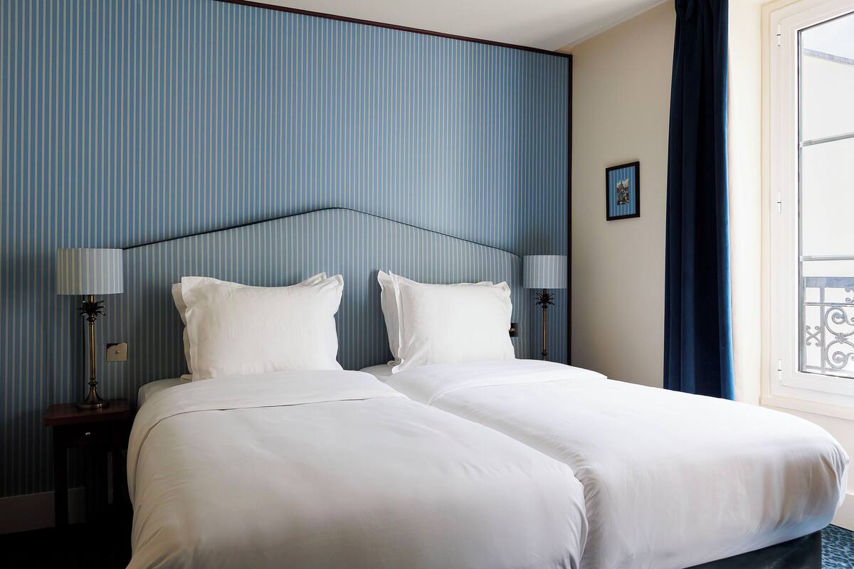 A double bed with white linens, a nightstand with a lamp, and a window with a curtain.