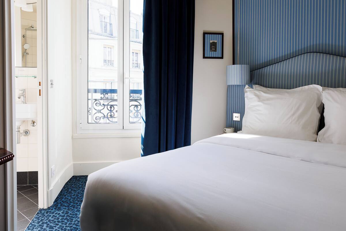 A hotel room with a bed, a window with a view of buildings, a bathroom with a shower, and a blue patterned floor.