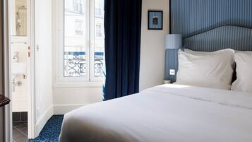 A hotel room with a bed, a window with a view of buildings, a bathroom with a shower, and a blue patterned floor.