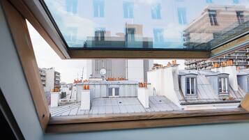 Une vue depuis une fenêtre de toit donnant sur un paysage urbain avec de nombreux bâtiments, dont un imposant bâtiment blanc à l’architecture moderne et un bâtiment traditionnel doté d’un toit en pente.