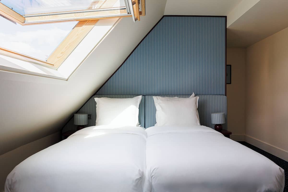 A bedroom with a skylight, a bed with white bedding, a nightstand with a lamp, and a striped wall.