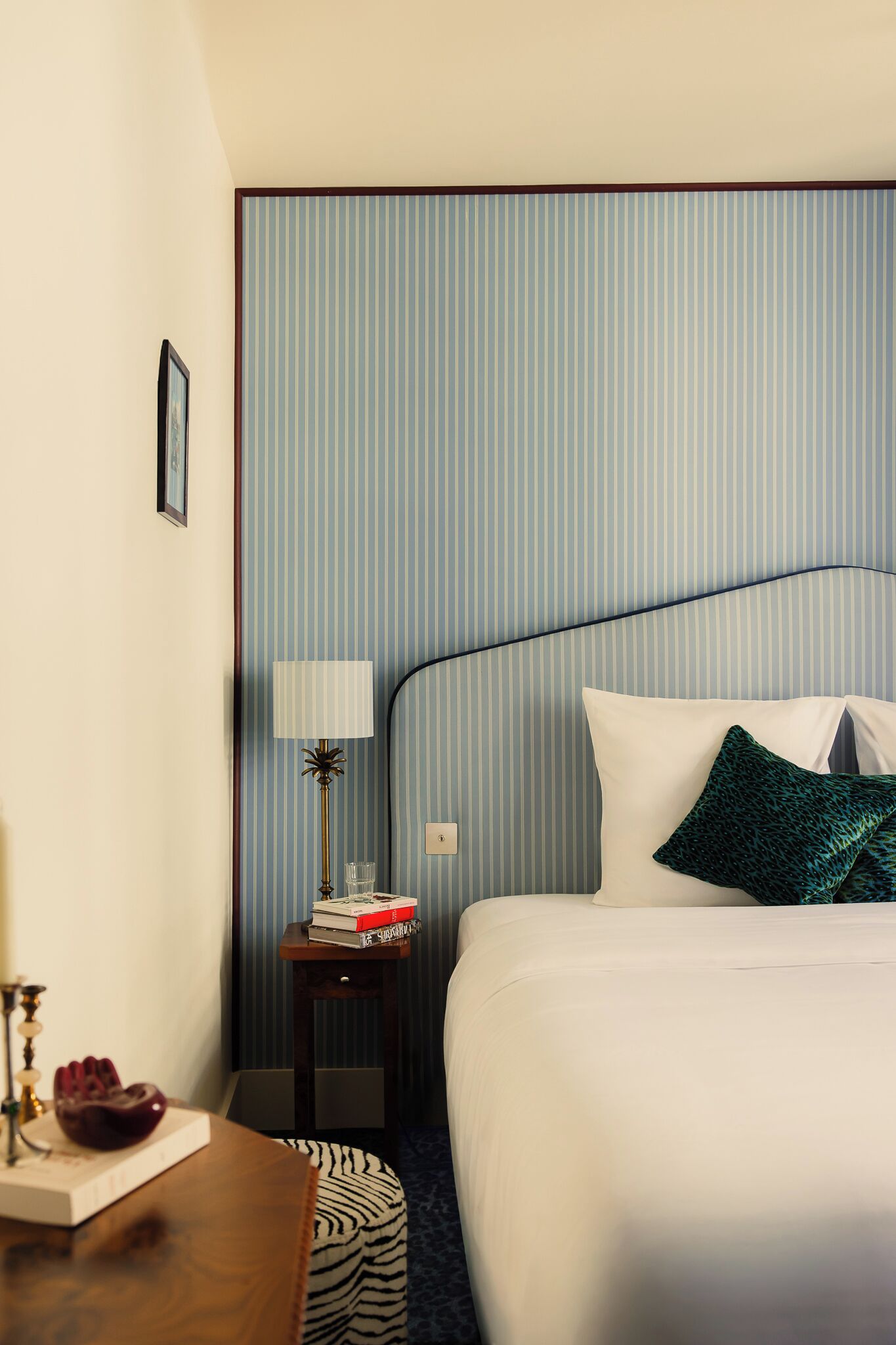 A hotel room with a bed, a nightstand with a lamp, a small table with a book and a decorative item, and a striped wall.