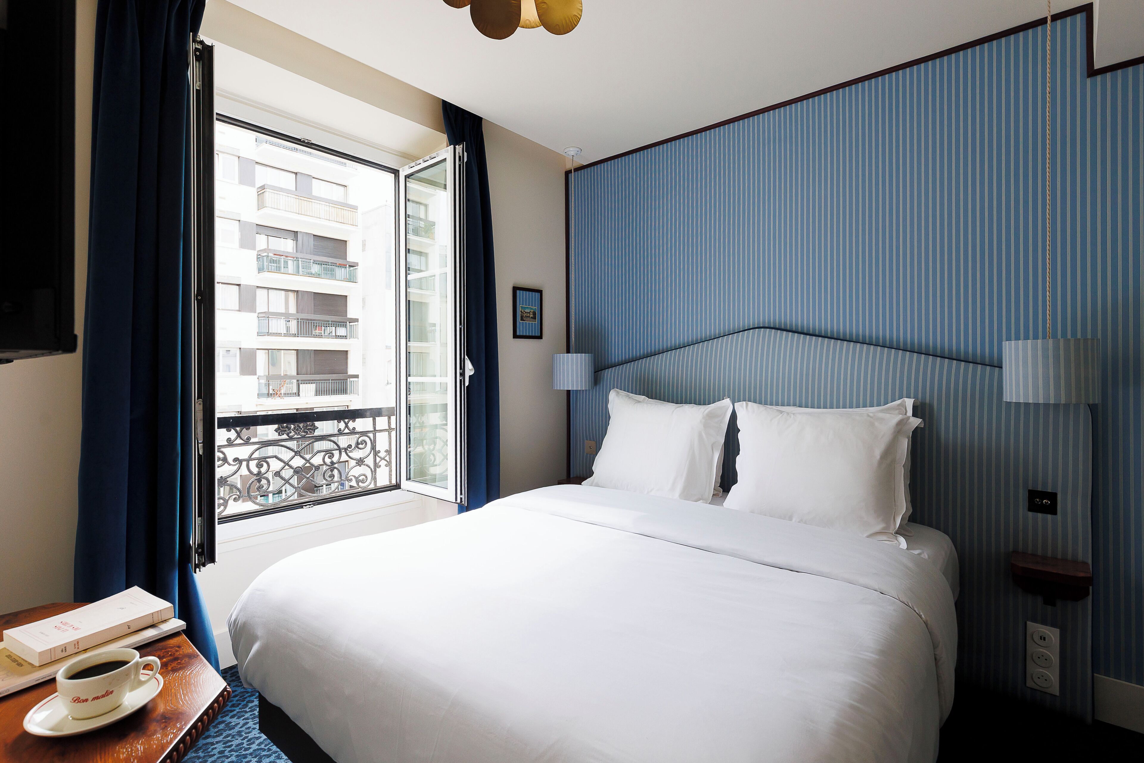 A hotel room with a bed, a window with curtains, a bedside table with a cup and a book, and a wall with a striped pattern.