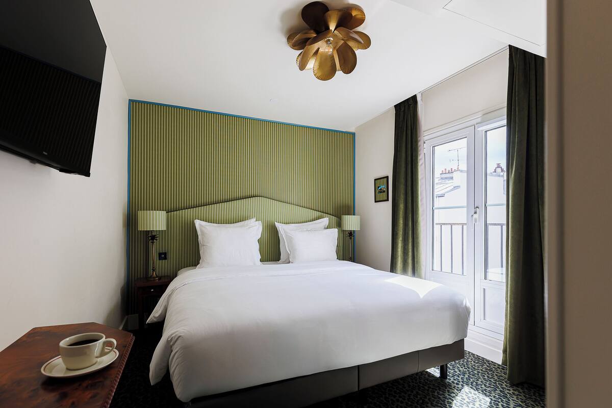 A hotel room with a bed, a coffee cup on a table, a TV mounted on the wall, and a view of the outside through a window.