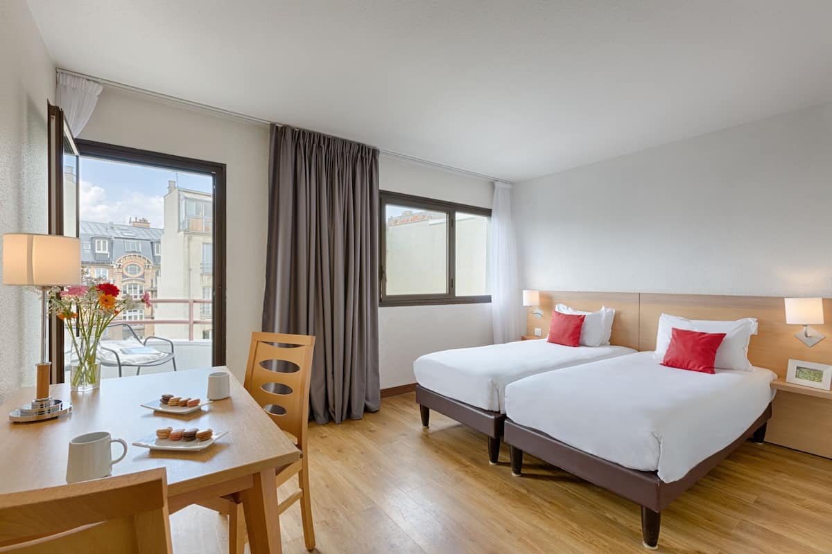 Une chambre d’hôtel avec deux lits, une table à manger, des chaises, une fenêtre avec des rideaux et une vue sur les bâtiments environnants.