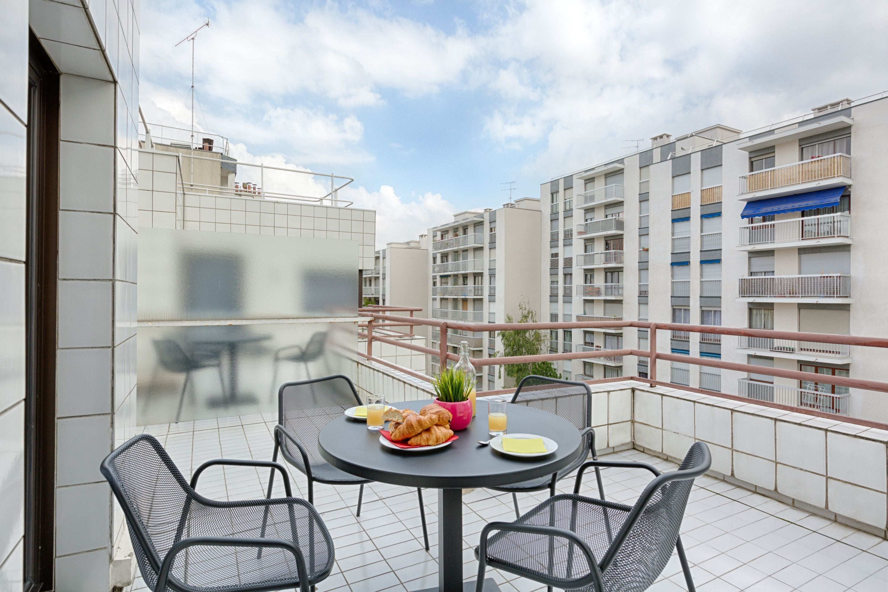Un balcon avec une table ronde, quatre chaises, une bouteille d’eau, un verre de jus d’orange et une assiette de croissants.