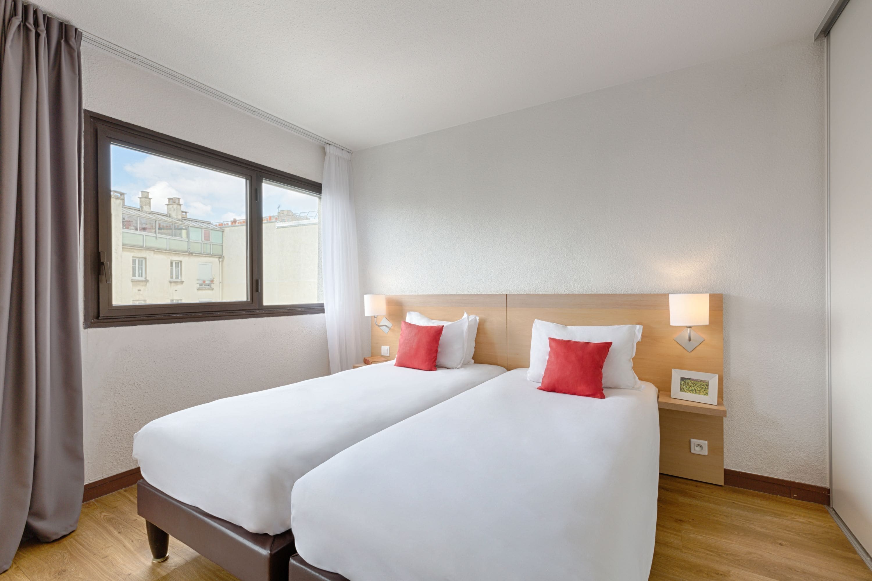 A hotel room with two beds, a window with curtains, a nightstand with a lamp, and a wall-mounted picture.