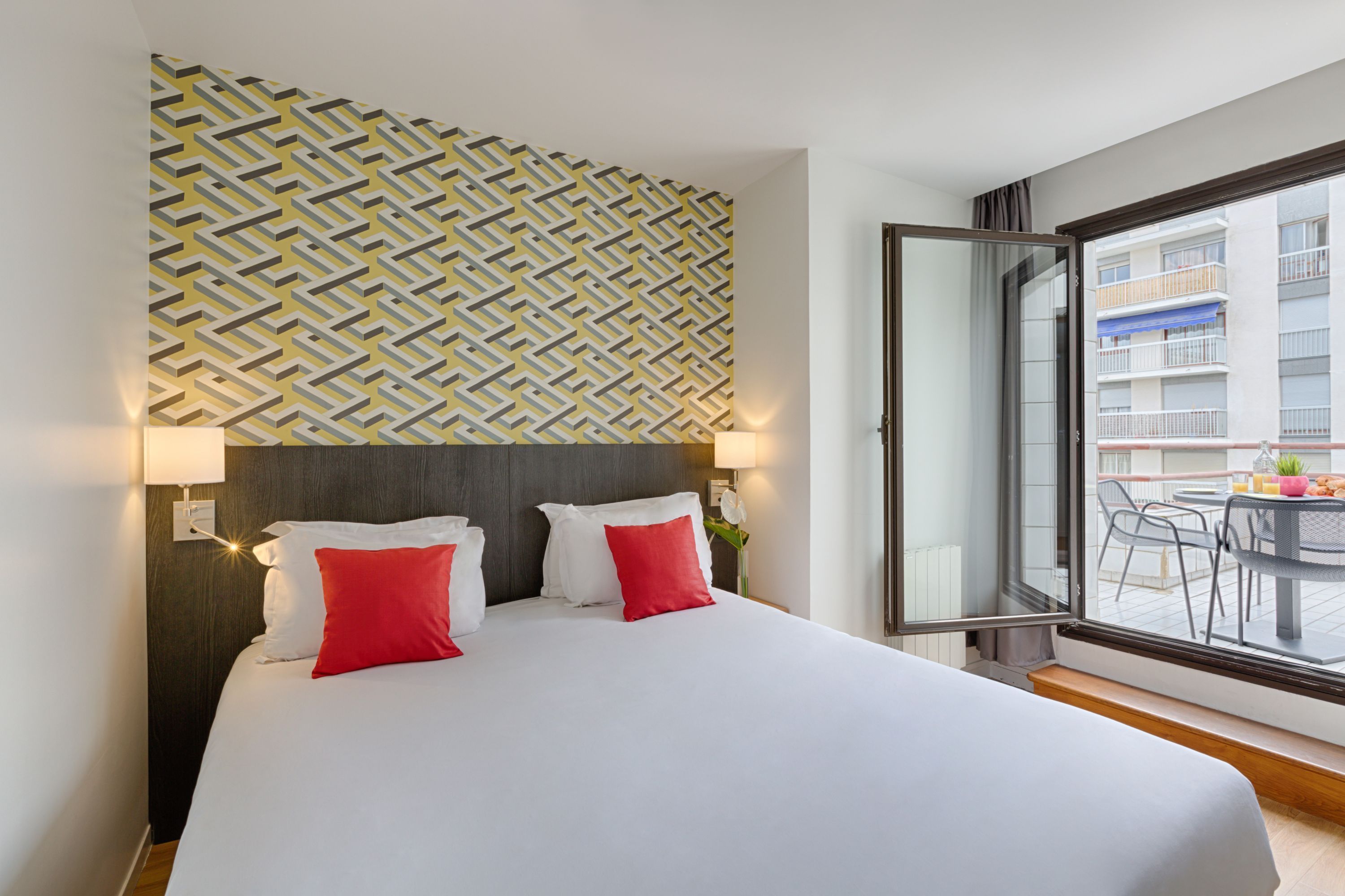 A hotel room with a large bed, two red pillows, a geometric patterned wall, and a balcony with outdoor furniture.