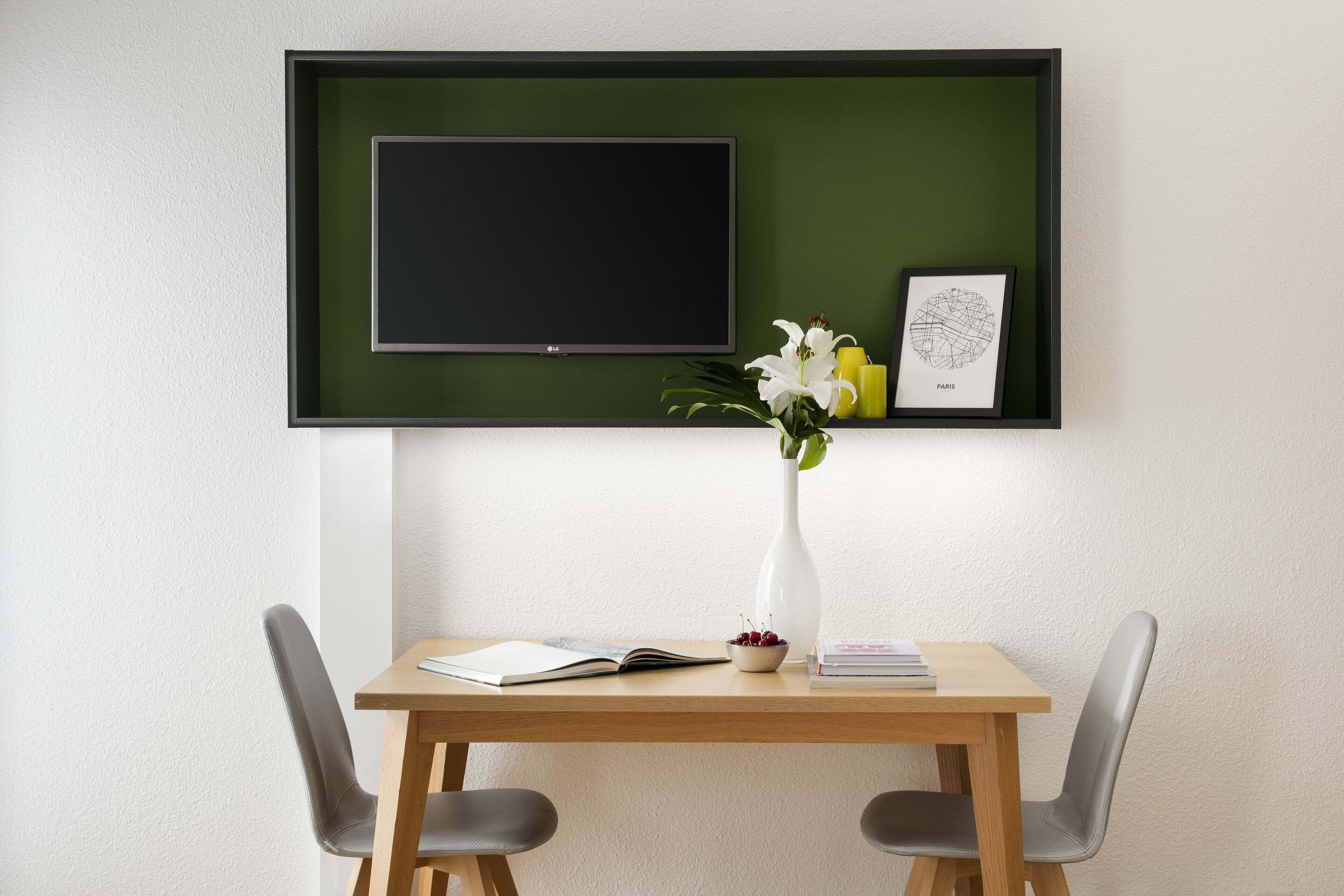 A modern room with a flat-screen TV mounted on a green wall, a wooden table with two chairs, a vase with flowers, and a framed map of Paris.