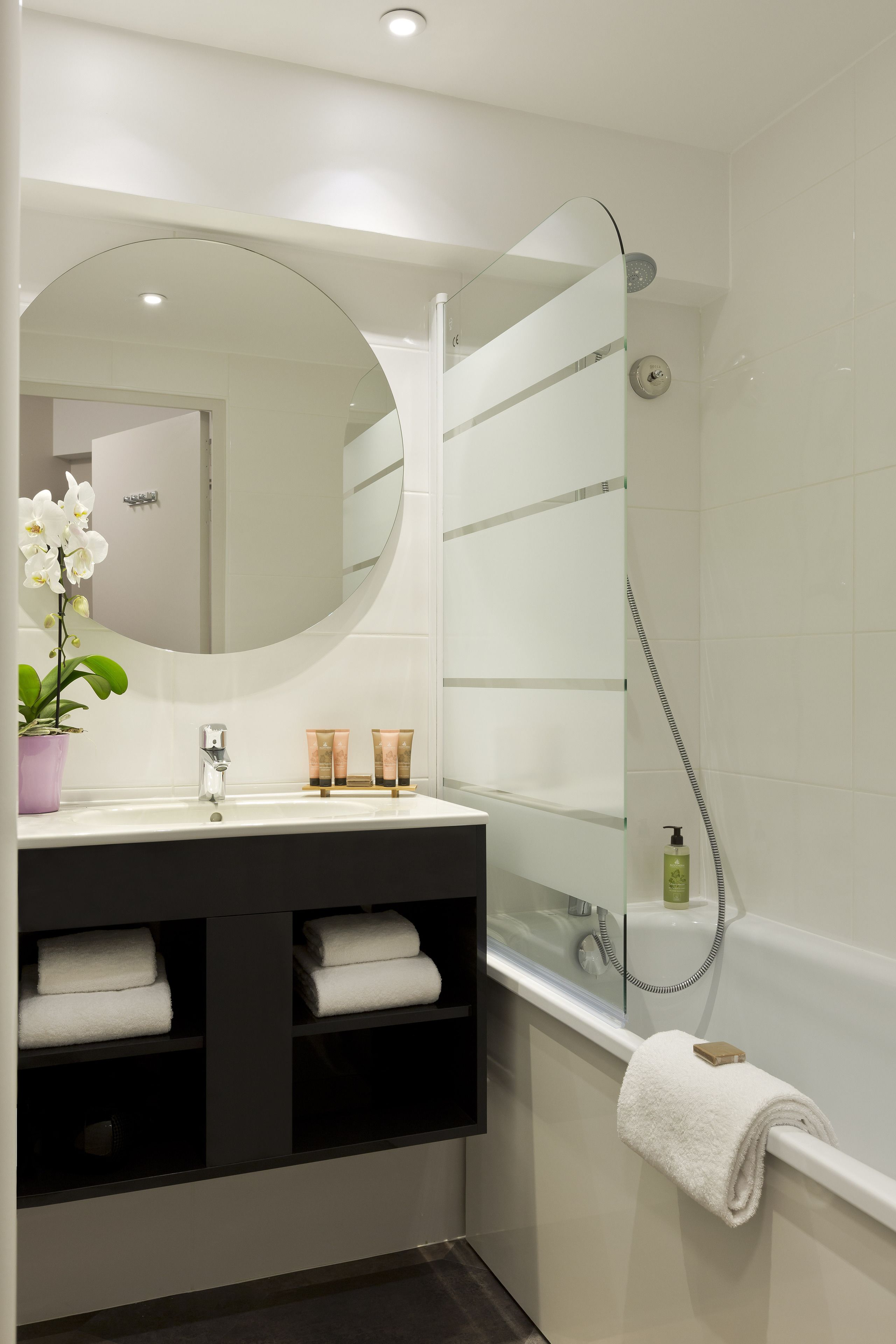 A modern bathroom with a round mirror, a sink with two towel racks, a glass shower enclosure, and a potted orchid.