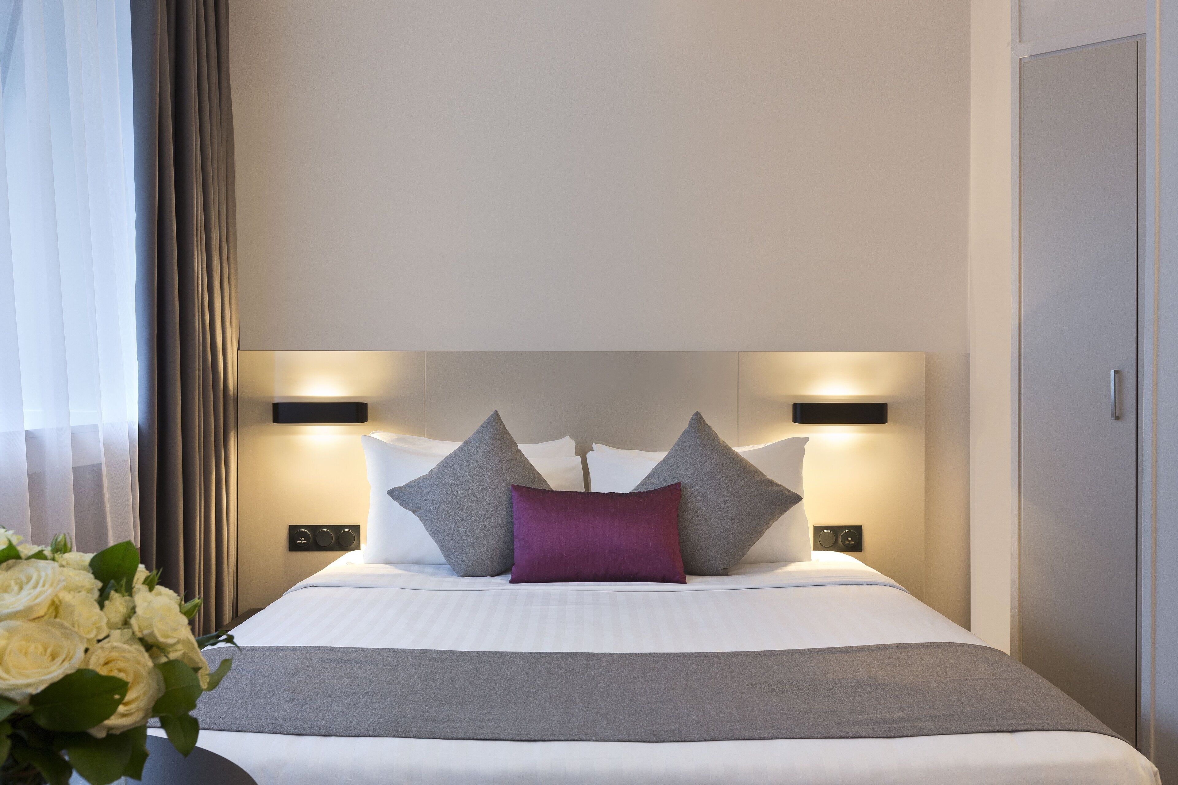 A neatly made bed with a grey and purple pillow arrangement, a bedside table with a lamp, and a wall-mounted light fixture.