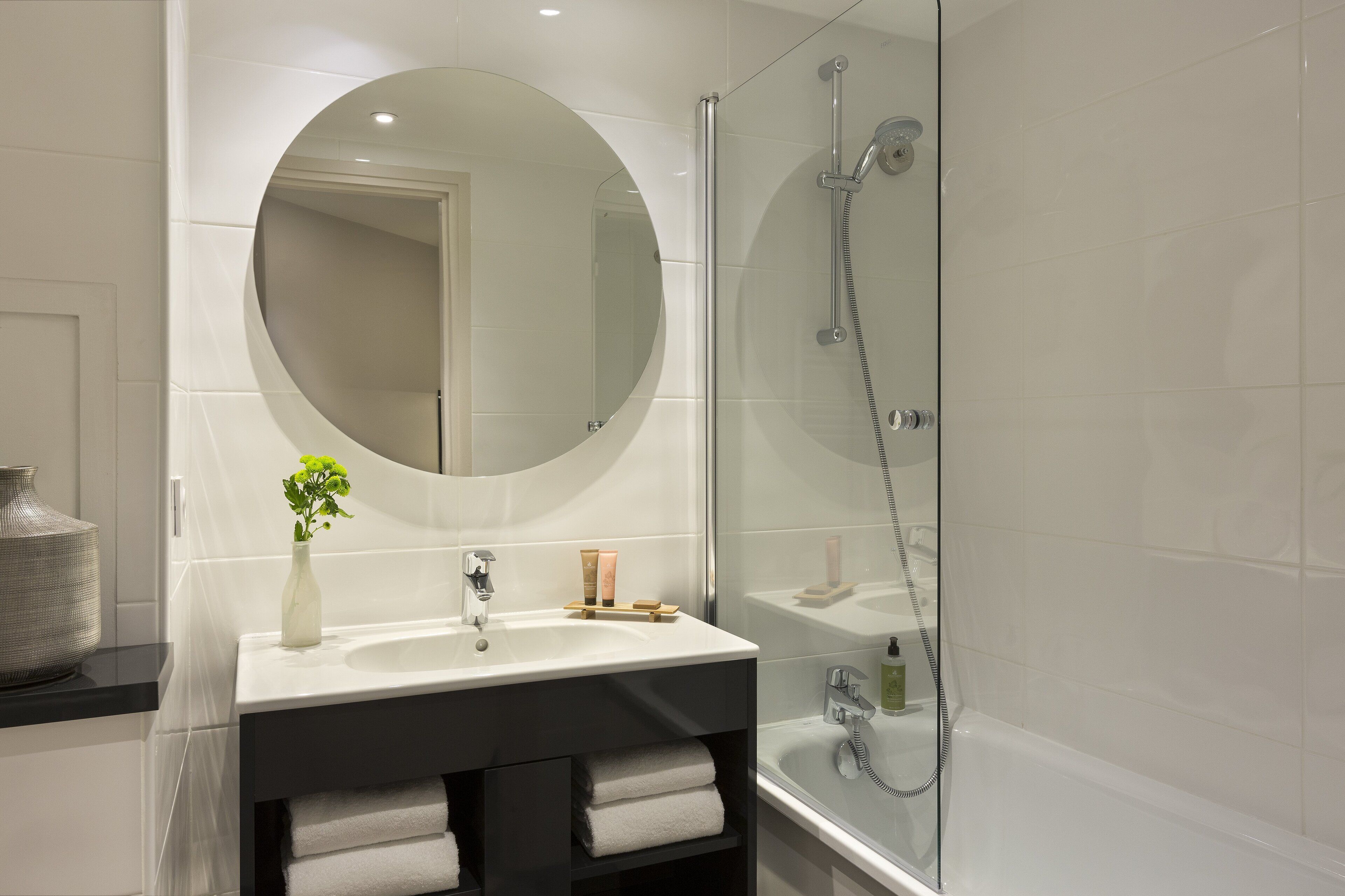 A modern bathroom with a round mirror, a white sink, and a glass-enclosed shower.