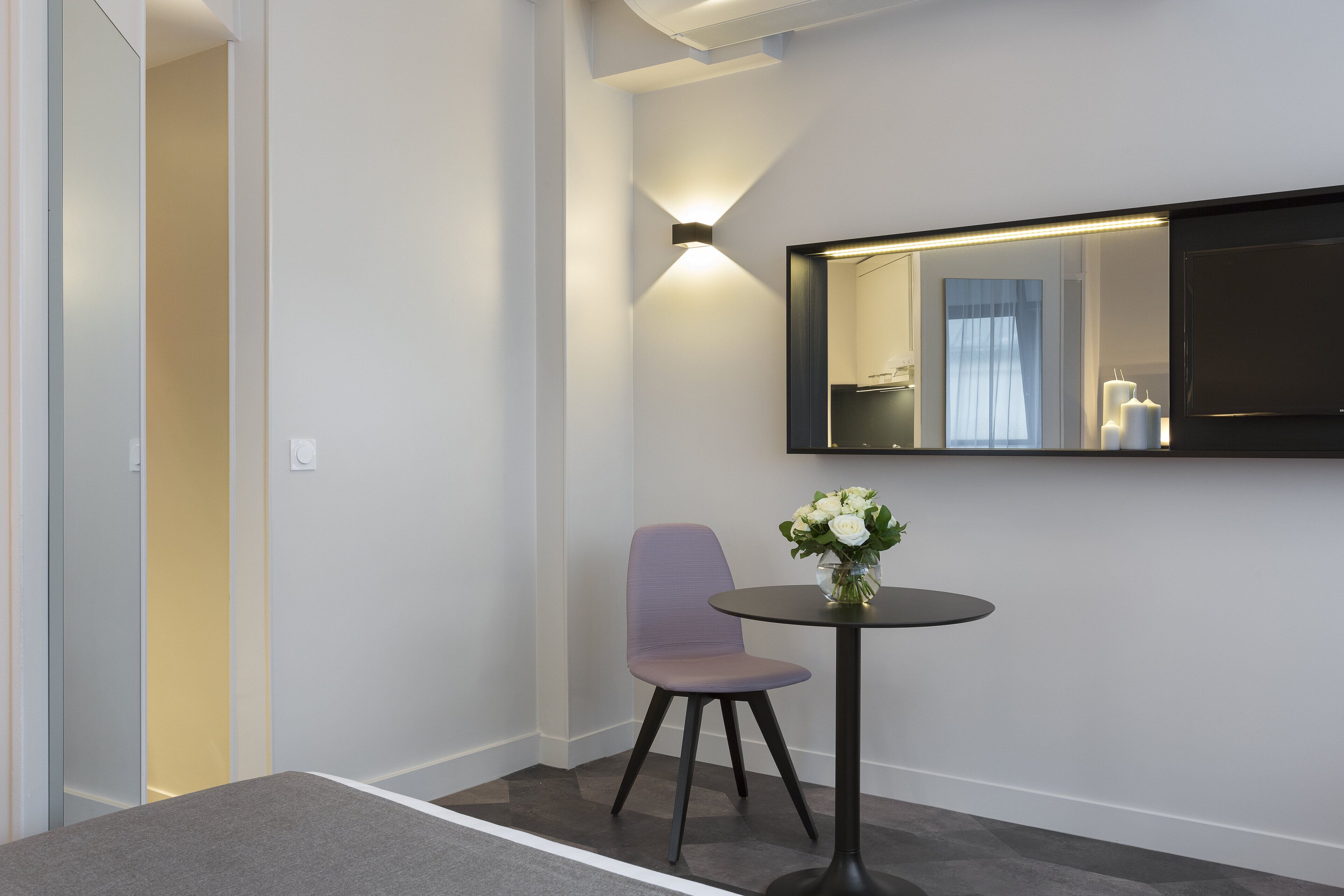 Une chambre d’hôtel moderne au design minimaliste, comprenant un seul fauteuil violet, une petite table ronde avec un vase de fleurs et un grand miroir mural.