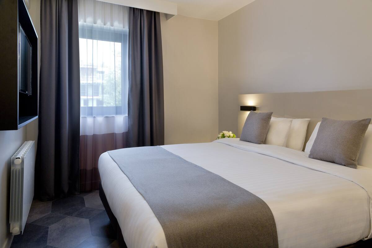 A hotel room with a large bed, a flat-screen TV, a bedside table with a lamp, and a window with curtains.