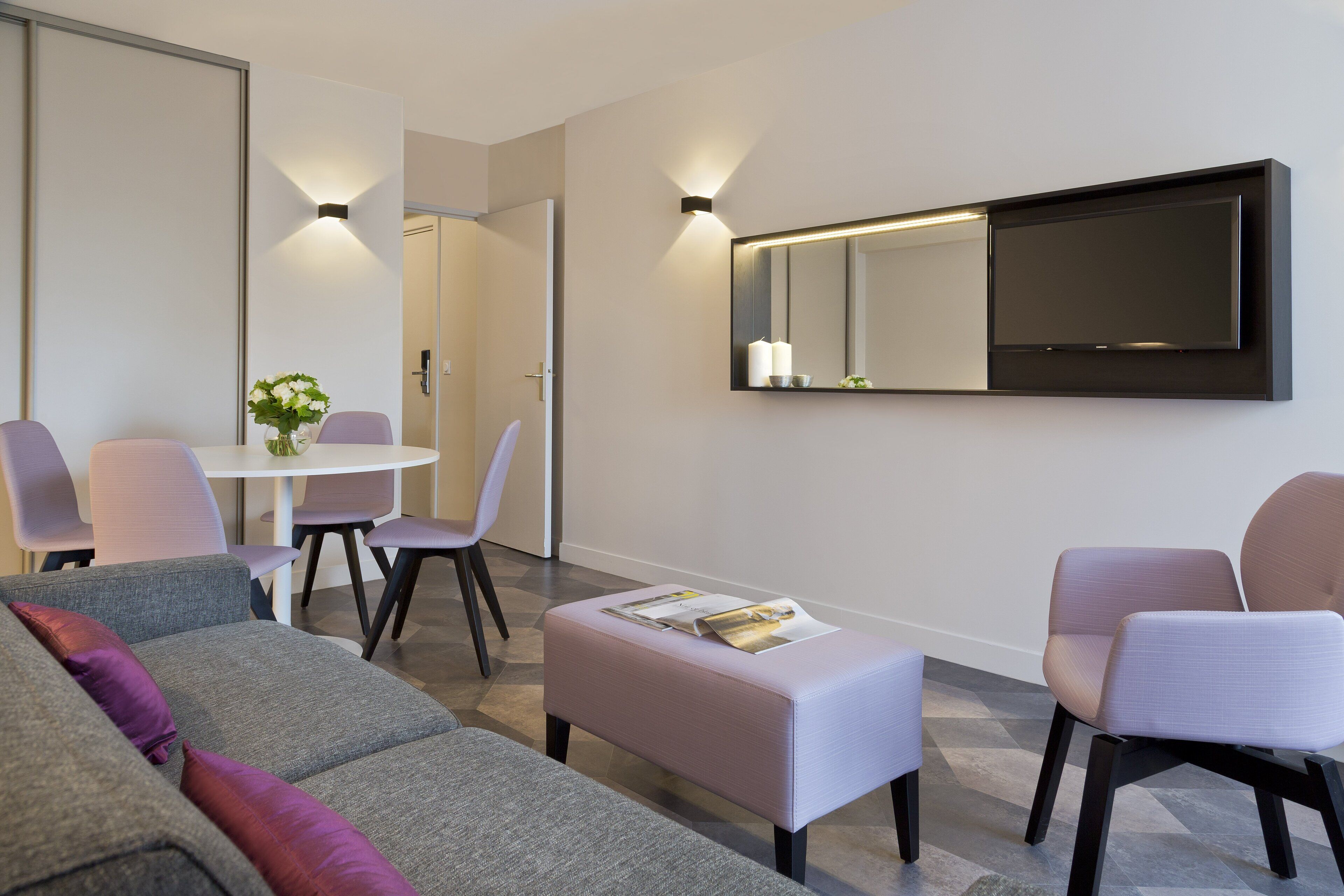 Una sala de estar moderna con un sofá gris, un reposapiés blanco y una mesa de centro. Se observa una zona de comedor con una mesa y sillas blancas. Un espejo grande y un televisor de pantalla plana están montados en la pared.