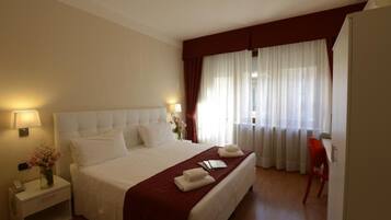 Une chambre d’hôtel avec un grand lit, une table de chevet avec une lampe, un fauteuil rouge et une fenêtre avec des rideaux légers.