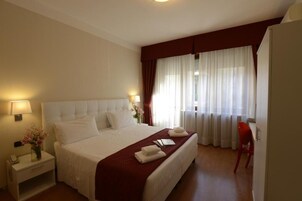 Camera d'albergo con un letto grande, un comodino con una lampada, una sedia rossa e una finestra con tende leggere.