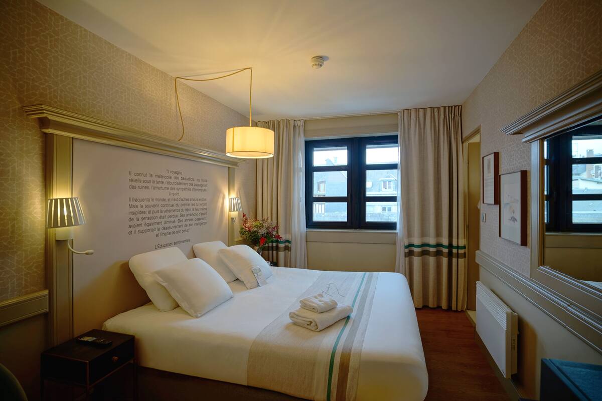 Une chambre d’hôtel avec un grand lit, une table de chevet, une lampe, une fenêtre avec des rideaux et un mur orné d’un cadre contenant un texte.
