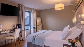 A hotel room with a bed, a desk, a chair, a TV, a window with curtains, and a bedside table with a vase.