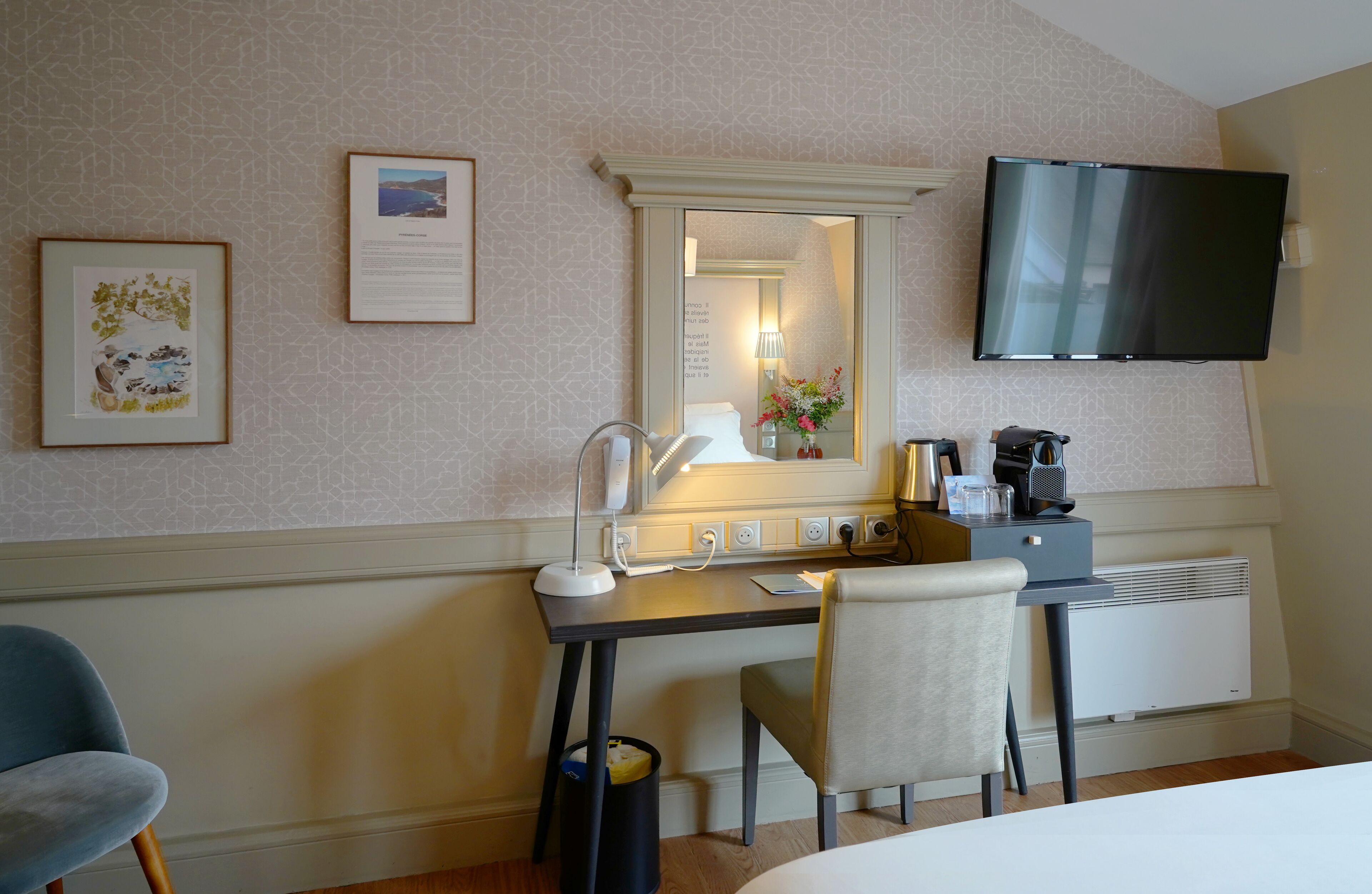 Ett hotellrum med ett skrivbord, en stol, en tv, inramade bilder och en spegel.