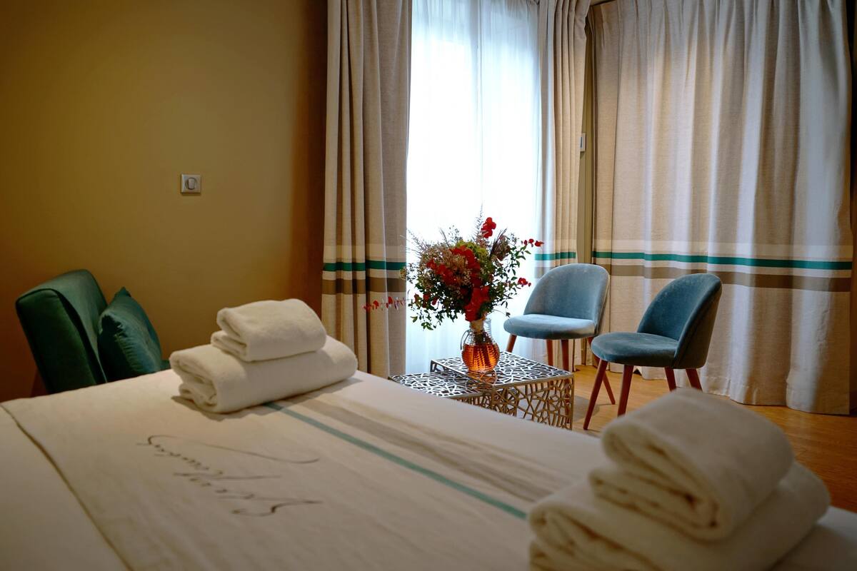 Une chambre d’hôtel avec un lit, deux chaises, une petite table ornée d’un vase de fleurs et une fenêtre avec des rideaux.