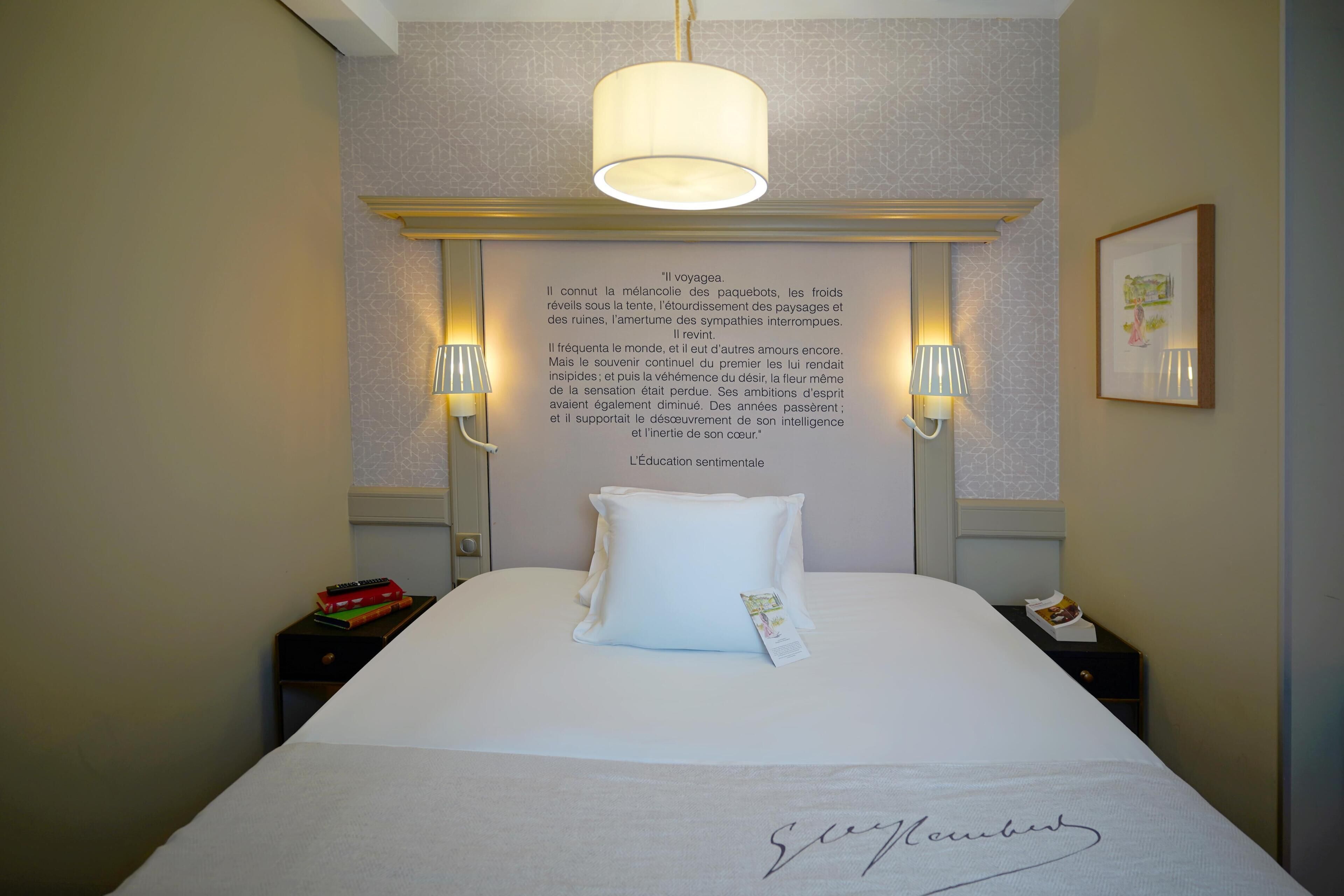 A neatly made bed with a pillow, a bedside table with a book, and a wall with a framed quote.