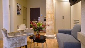 Een moderne lobby met een grijze bank, twee fauteuils, een ronde tafel met een bloevas en een hoge zuil.
