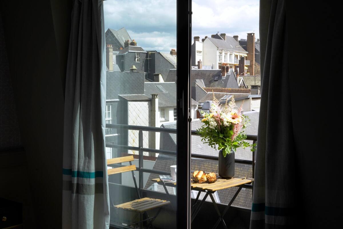 Une pièce avec vue sur le paysage urbain, visible à travers une fenêtre, une table en bois ornée d’un vase de fleurs et une chaise.