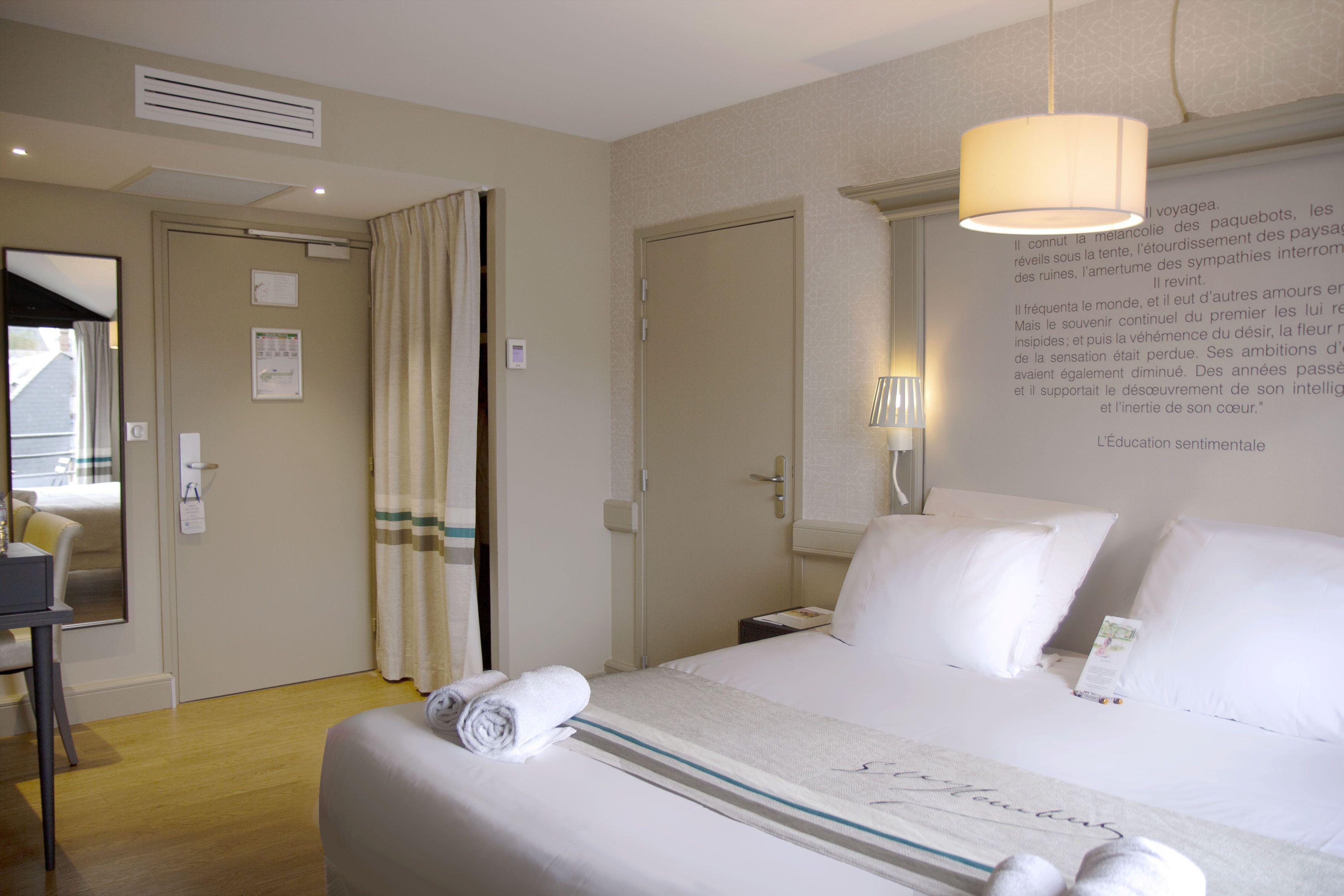 A hotel room with a neatly made bed, a desk, and a wall-mounted lamp.