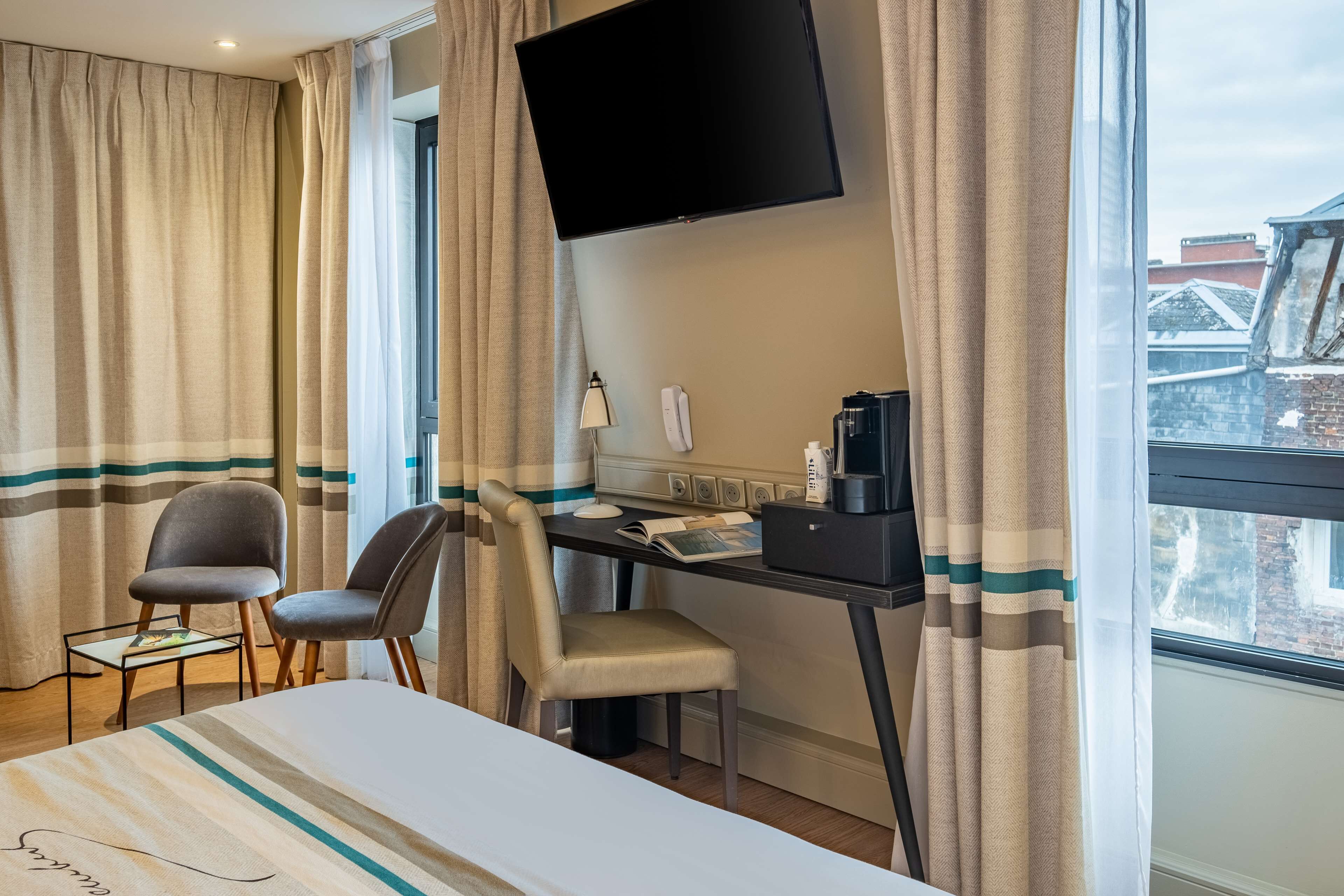 A hotel room with a bed, a desk with a computer, a chair, a television mounted on the wall, and a window with curtains.
