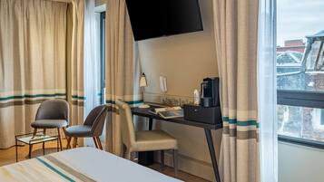 A hotel room with a bed, a desk with a computer, a chair, a television mounted on the wall, and a window with curtains.
