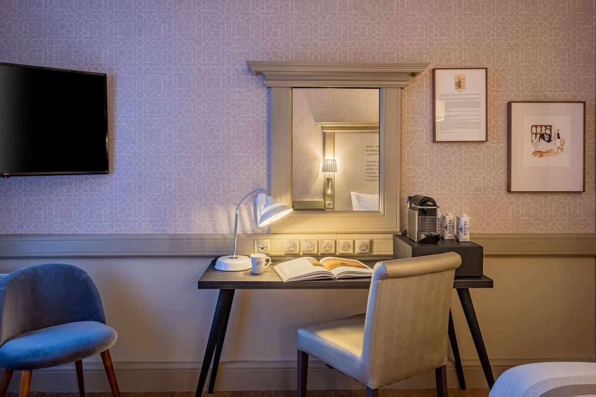 Une chambre d’hôtel équipée d’un bureau, de deux chaises, d’un téléviseur à écran plat, d’un miroir, d’œuvres d’art encadrées et d’une machine à café.