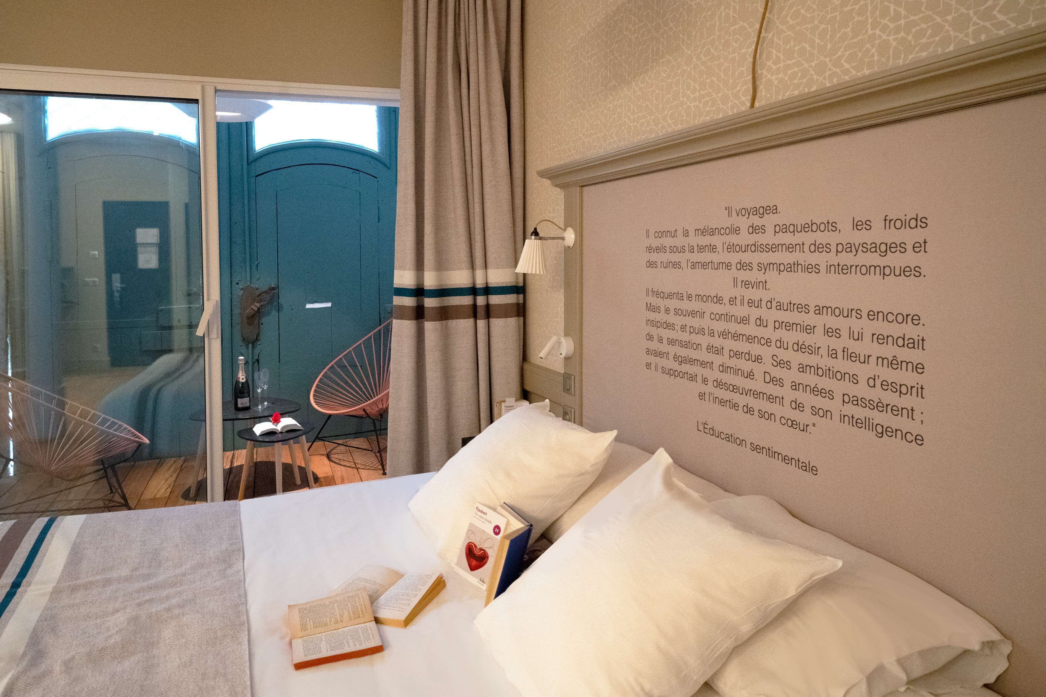 A hotel room with a bed, a bedside table with books, a wall with a quote, and a view of the outside.