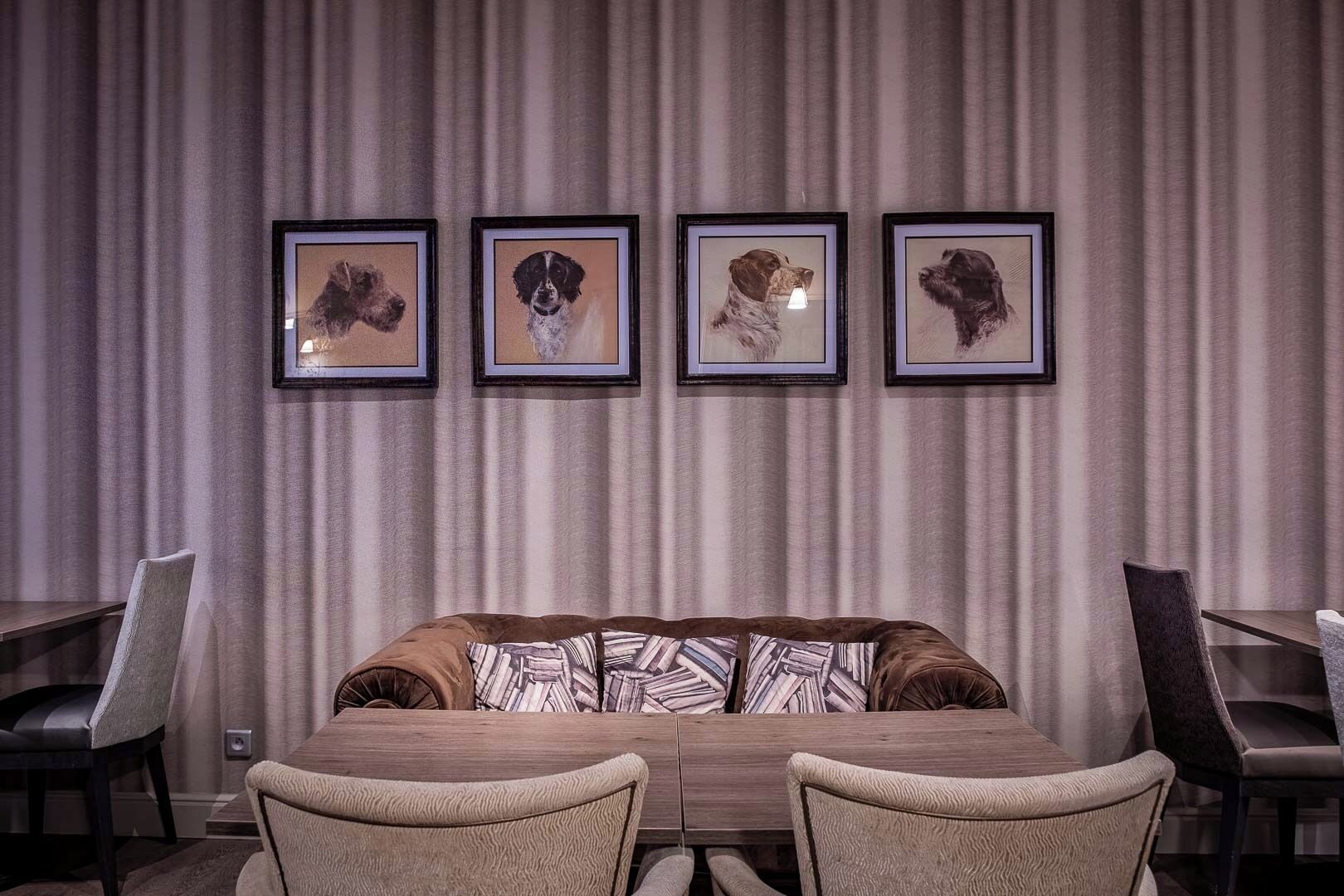 A dining area with a long table, four framed dog portraits on the wall, and a patterned throw on the sofa.