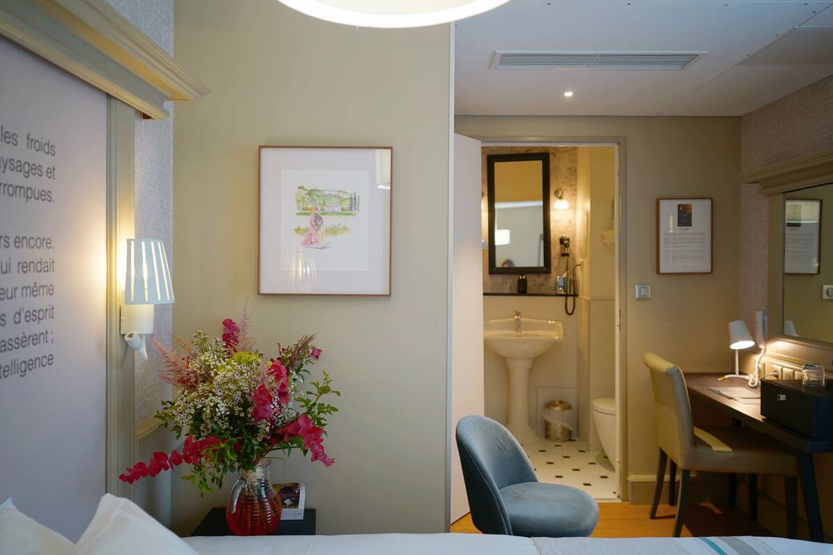 Une chambre d’hôtel comprenant un lit, un bureau, une chaise, une salle de bain avec un lavabo et un miroir, et un tableau encadré au mur.