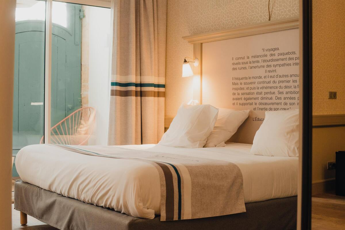 Un lit bien fait, avec du linge de lit blanc et une couverture à rayures, une lampe de chevet et un mur orné d’un tableau.
