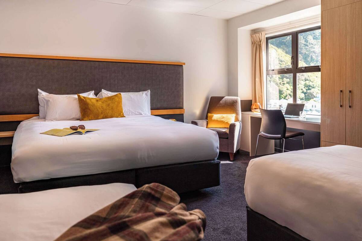 A hotel room with two beds, a desk, a chair, and a large window with a view of greenery.