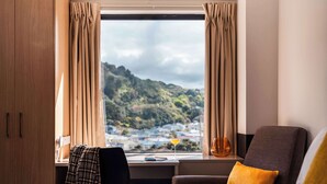 Ein Hotelzimmer mit einem Bett, einem Schreibtisch und einem Stuhl. Es gibt ein Fenster mit Vorhängen und einen Blick auf eine hügelige Landschaft.