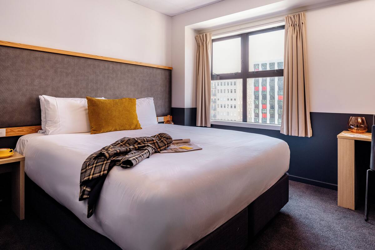 A hotel room with a large bed, a nightstand, a window with curtains, and a view of buildings outside.