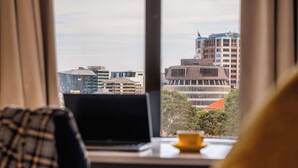 Ein Hotelzimmer mit einem Laptop auf dem Schreibtisch, einer Tasse am Fensterbrett und Blick auf eine Stadtlandschaft mit verschiedenen Gebäuden.