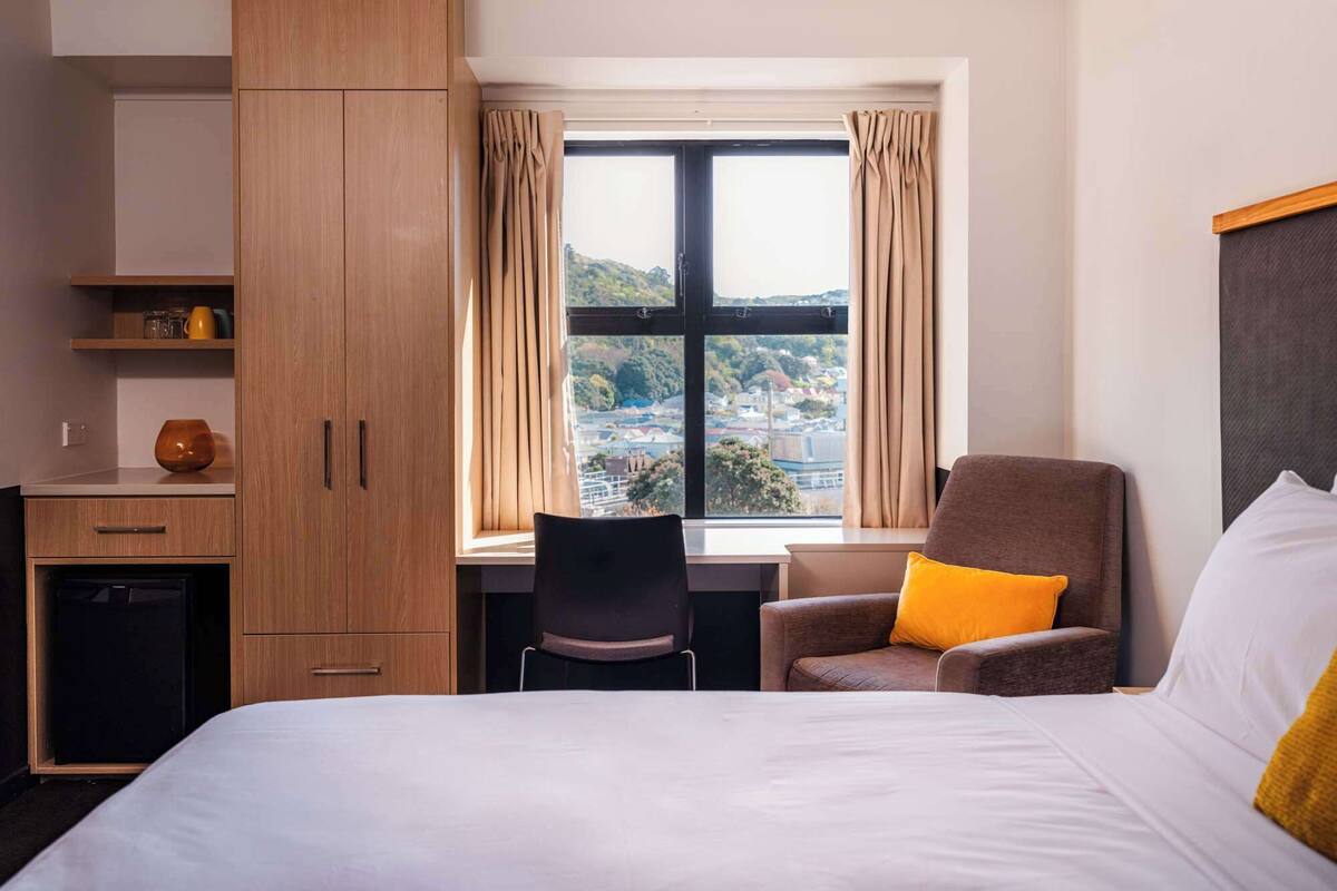 A hotel room with a bed, a desk, a chair, and a view of a cityscape through a large window.