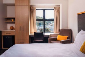 A hotel room with a bed, a desk, a chair, and a view of a cityscape through a large window.