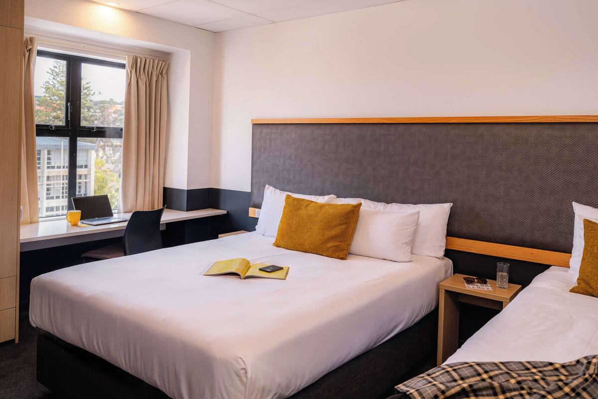 A hotel room with a large bed, a desk with a chair, a window with curtains, and a view of buildings outside.