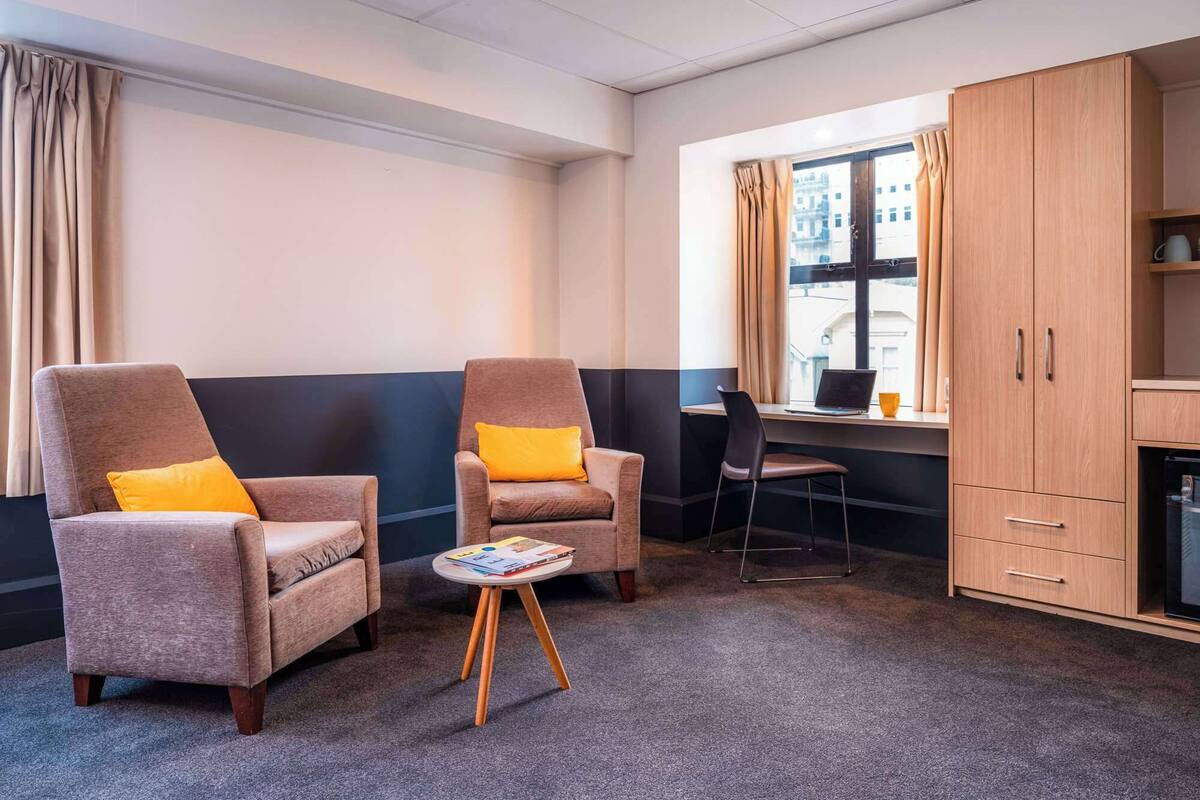 A hotel room with two armchairs, a small table, a desk with a chair, and a wardrobe.