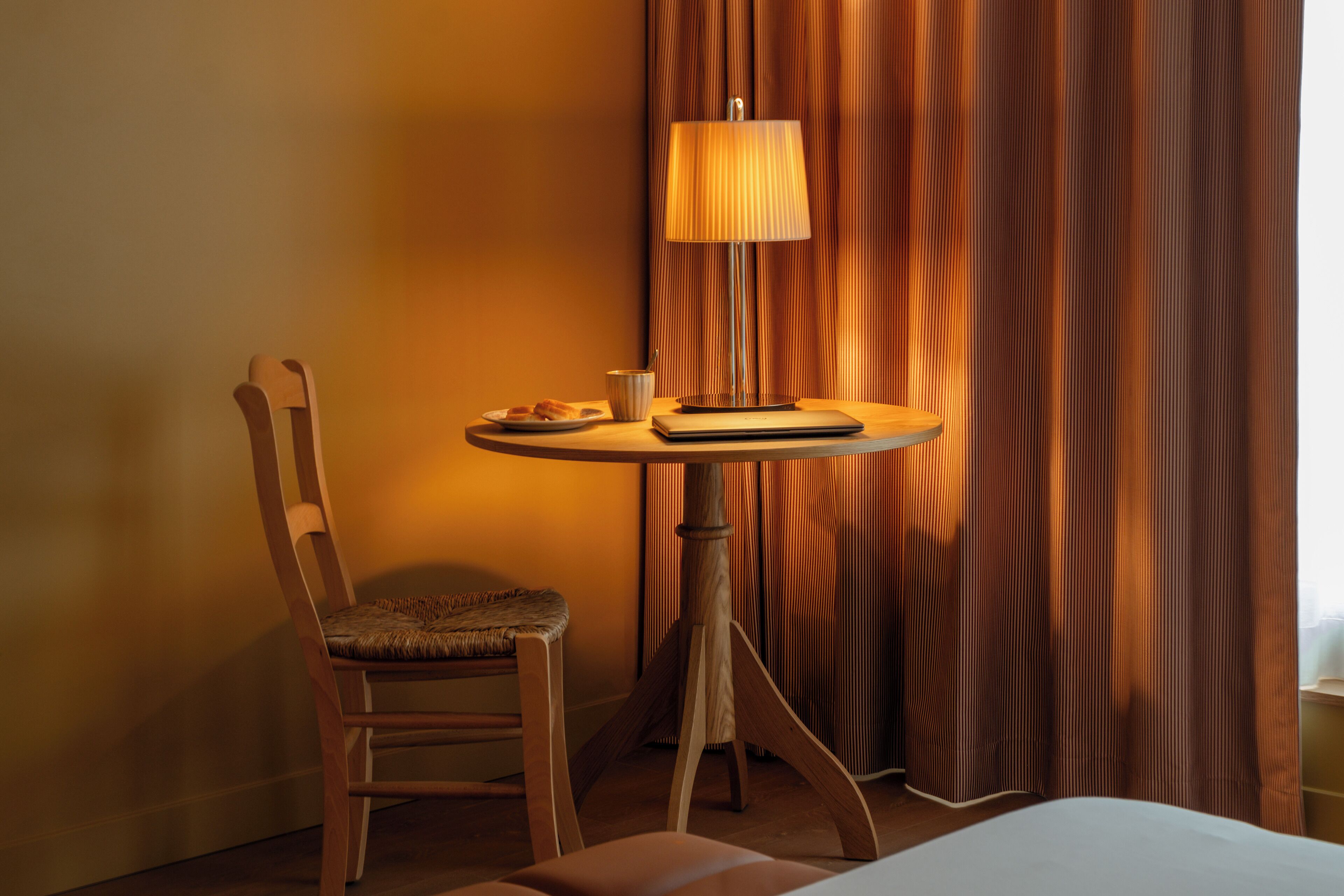A hotel room with a wooden chair, a small table with a lamp, a plate of croissants, and a book.
