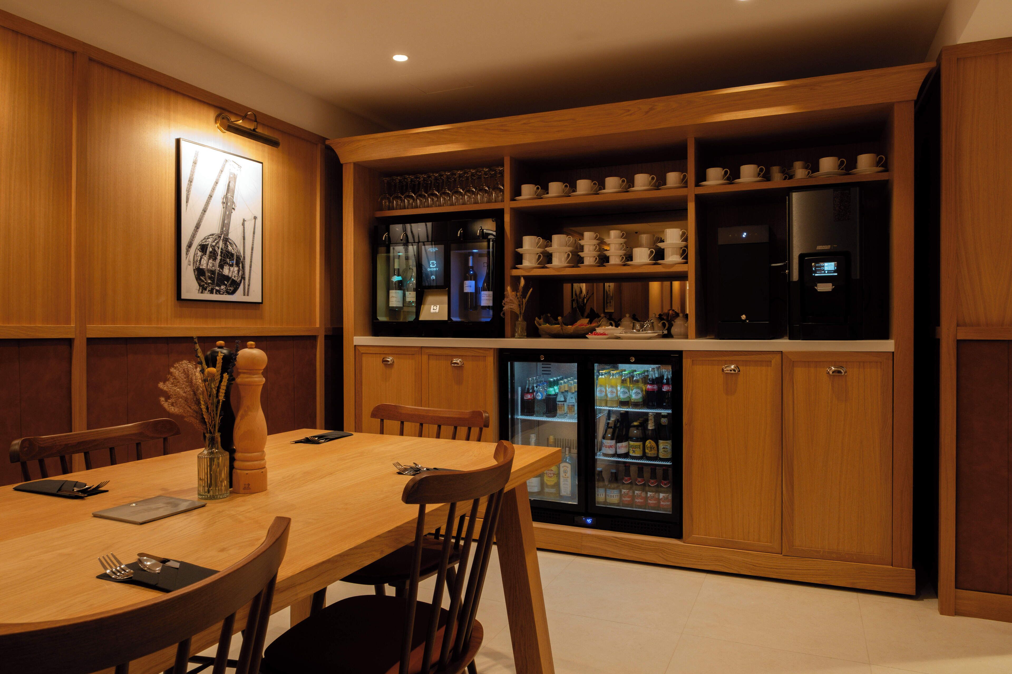 A dining area with a wooden table and chairs, a refrigerator, and shelves with cups and glasses.