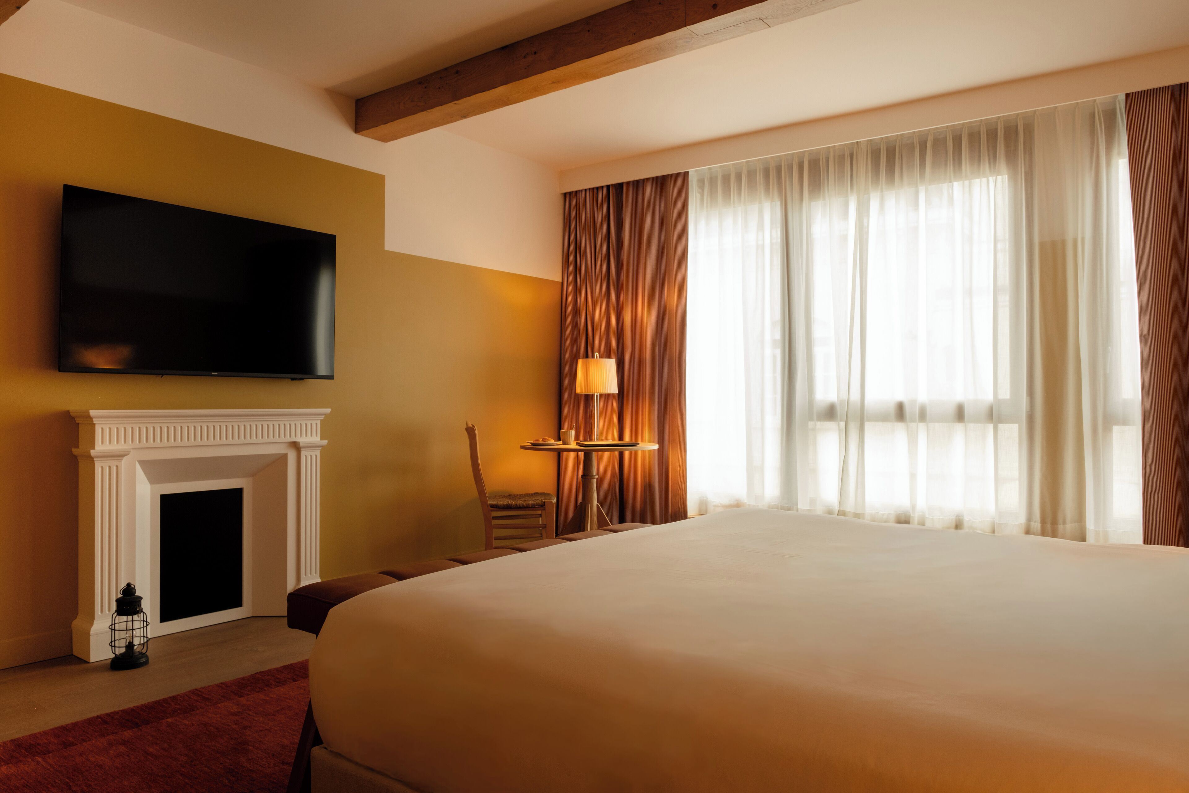 Une chambre d’hôtel avec un grand lit, un téléviseur à écran plat fixé au mur, une cheminée, une table avec une lampe et une fenêtre avec des rideaux.