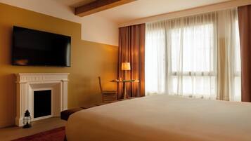 Une chambre d’hôtel avec un grand lit, un téléviseur à écran plat fixé au mur, une cheminée, une table avec une lampe et une fenêtre avec des rideaux.